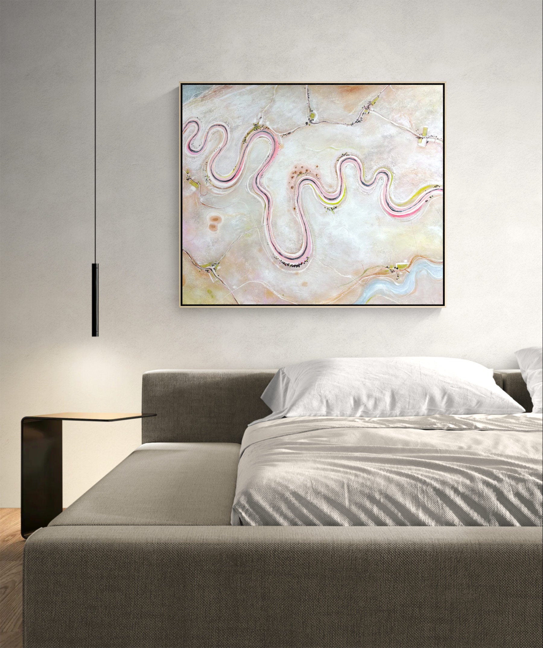 A square framed abstract wall art print hangs above a bed in a modern bedroom. The artwork features flowing, wavy lines in pink, yellow, and blue over a soft cream and beige background.