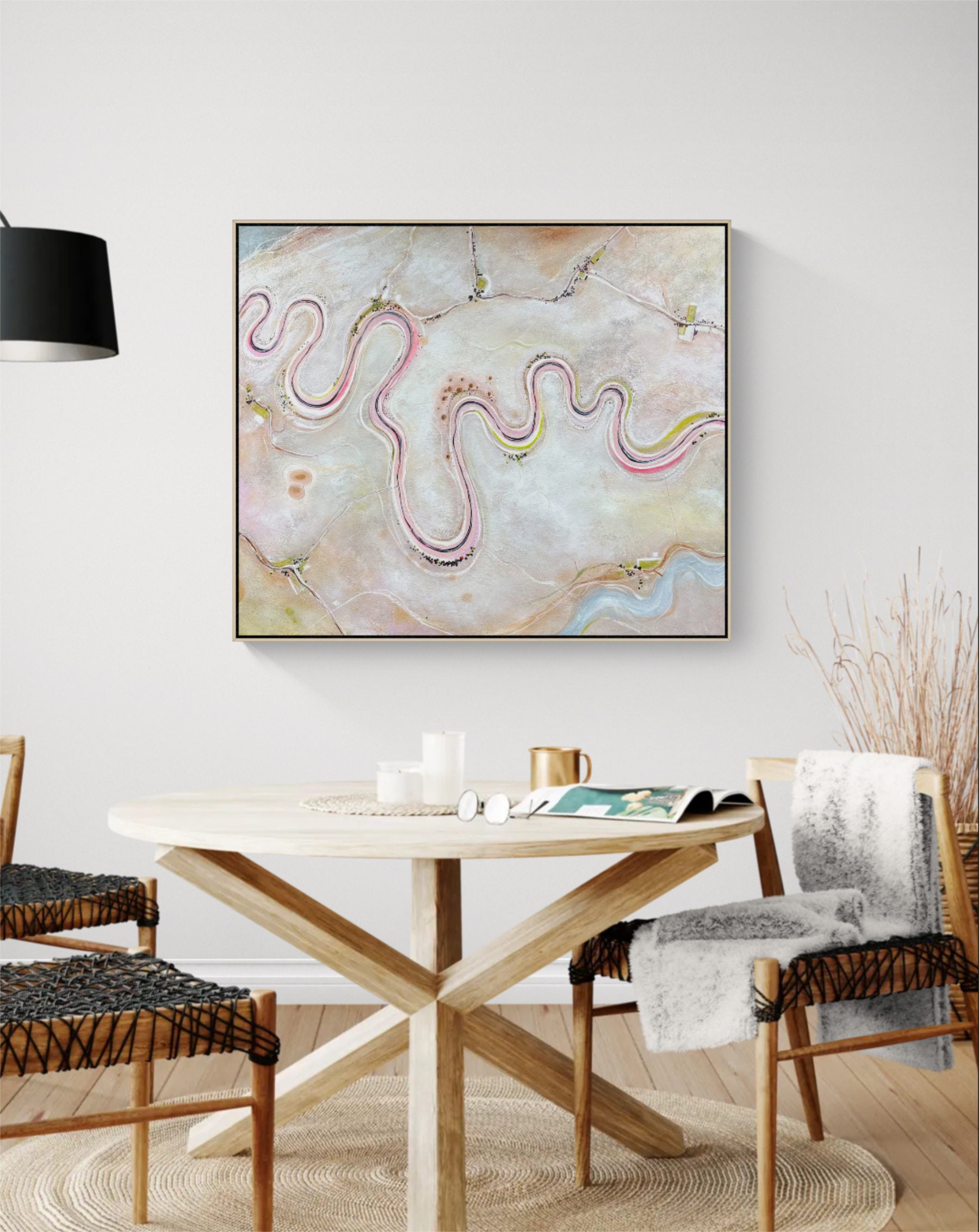 Abstract framed wall art hanging above a round wooden dining table in a bright, modern dining room. The artwork features flowing wavy lines in pink, gold, and blue over a soft beige and cream background.