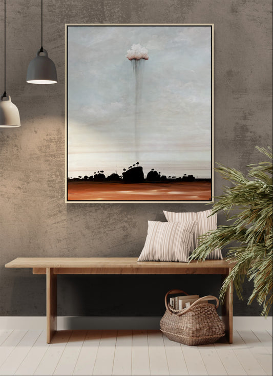 A framed landscape art original painting hangs on a textured gray wall above a wooden bench with striped pillows and a woven basket. The artwork depicts a dramatic cloud formation over a dark silhouetted landscape, with a muted blue-gray sky and a rust-brown horizon.