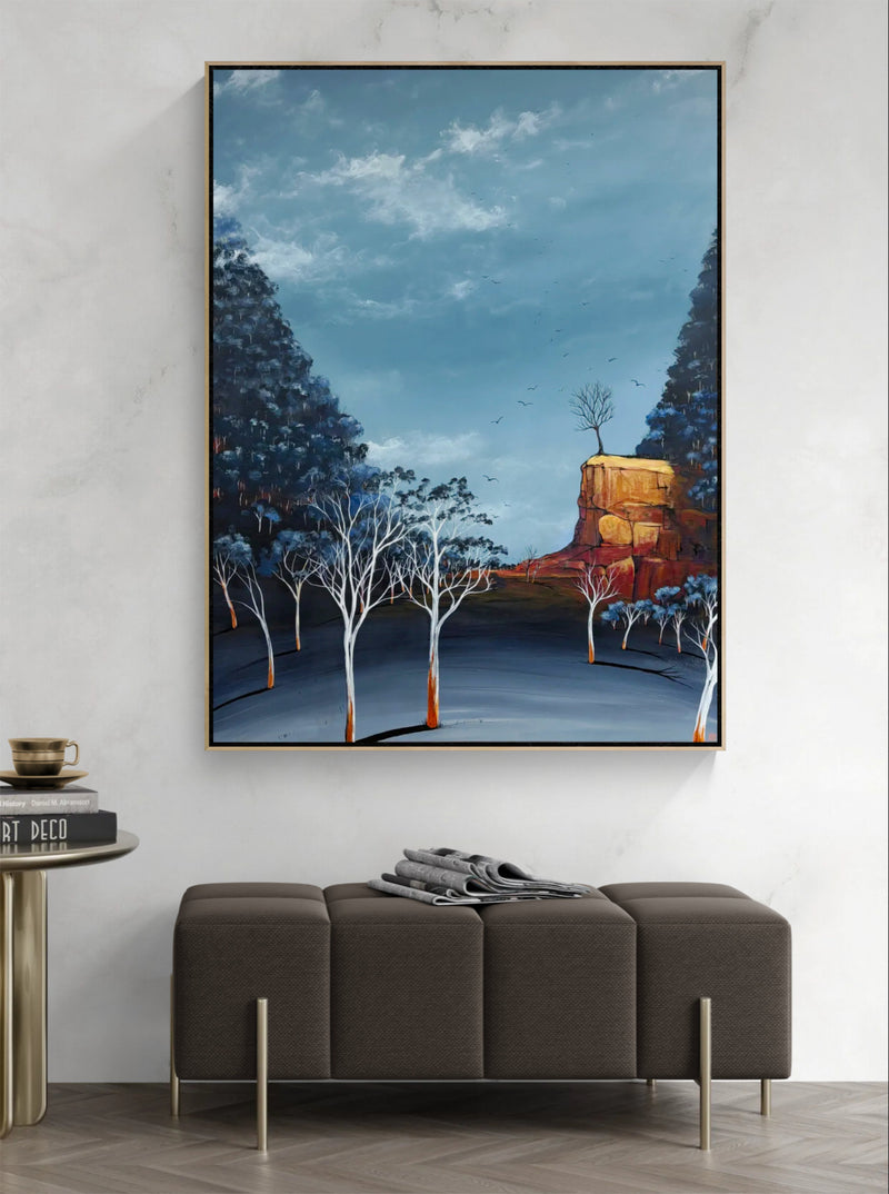 A framed landscape painting of a blue-toned forest with white-barked trees and a golden rock formation, displayed on a light gray wall above a brown bench in a modern interior setting.