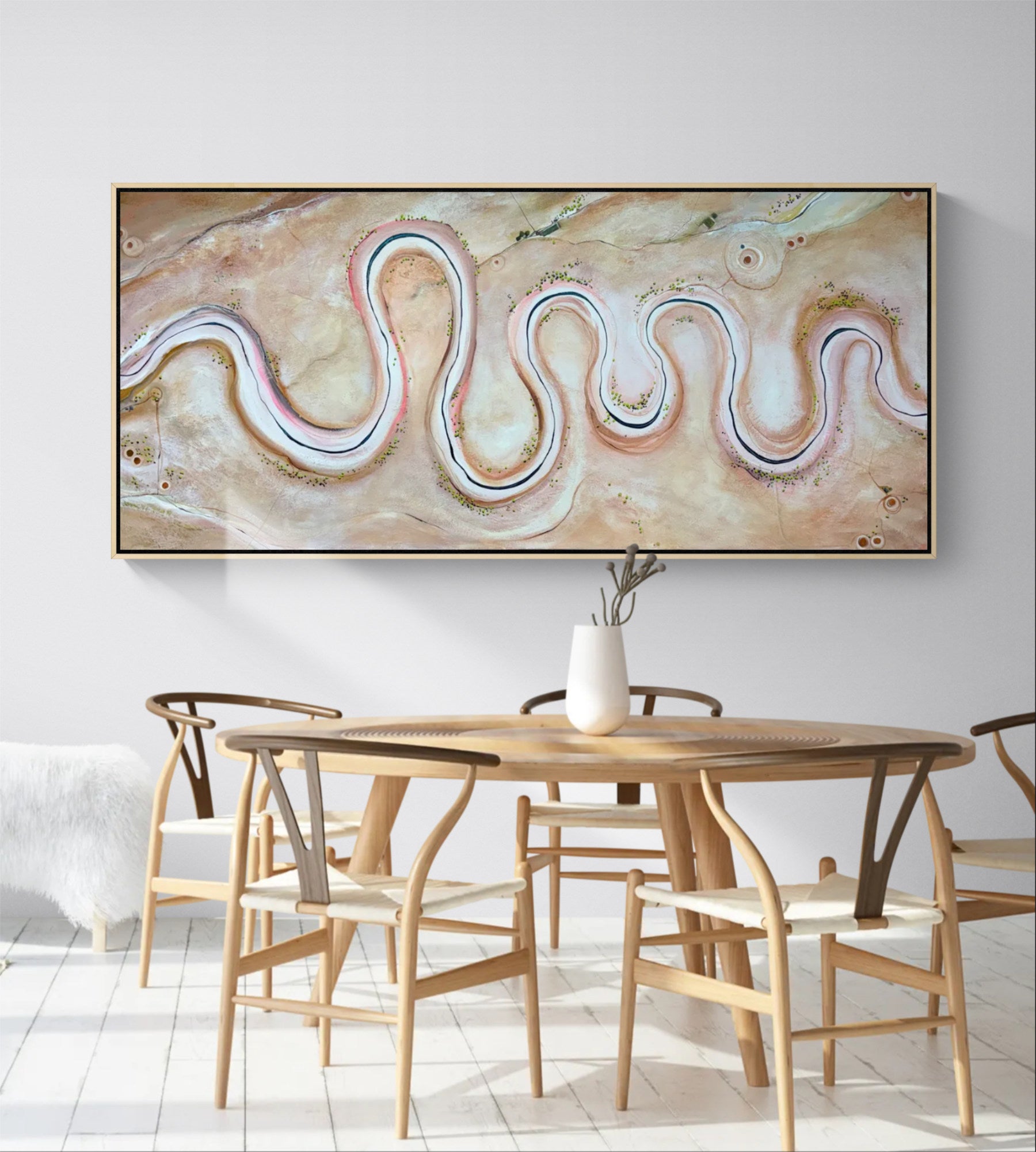 A large framed abstract wall art piece hangs above a wooden dining table and chairs in a bright, modern room. The artwork features flowing, wavy lines in cream, beige, blush pink, and soft blue tones.
