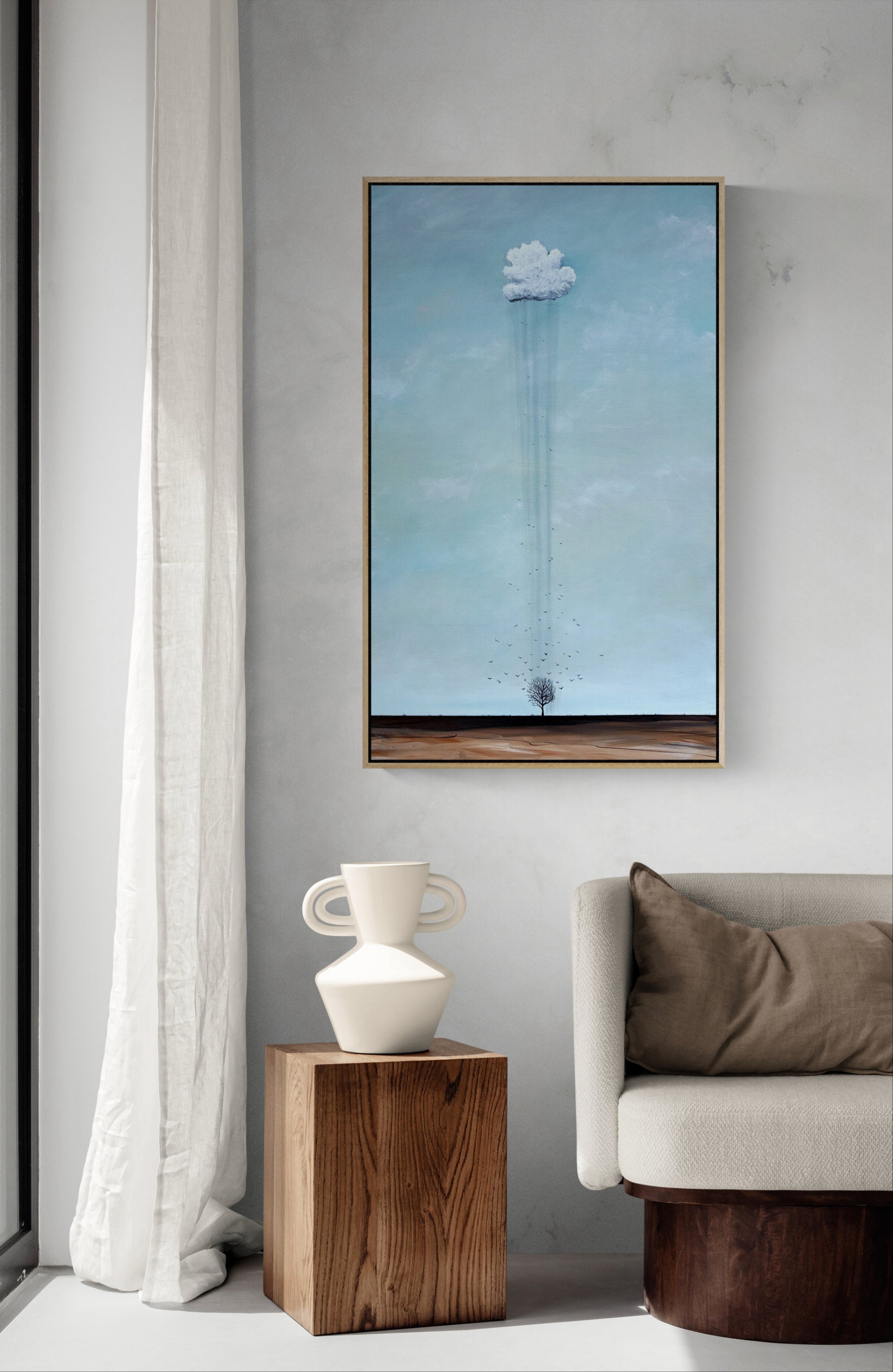 A vertical framed art print depicting a white cloud on a long stem against a light blue sky, displayed on a gray wall in a modern living room setting next to a white curtain, a wooden side table with a white vase, and a light gray sofa.