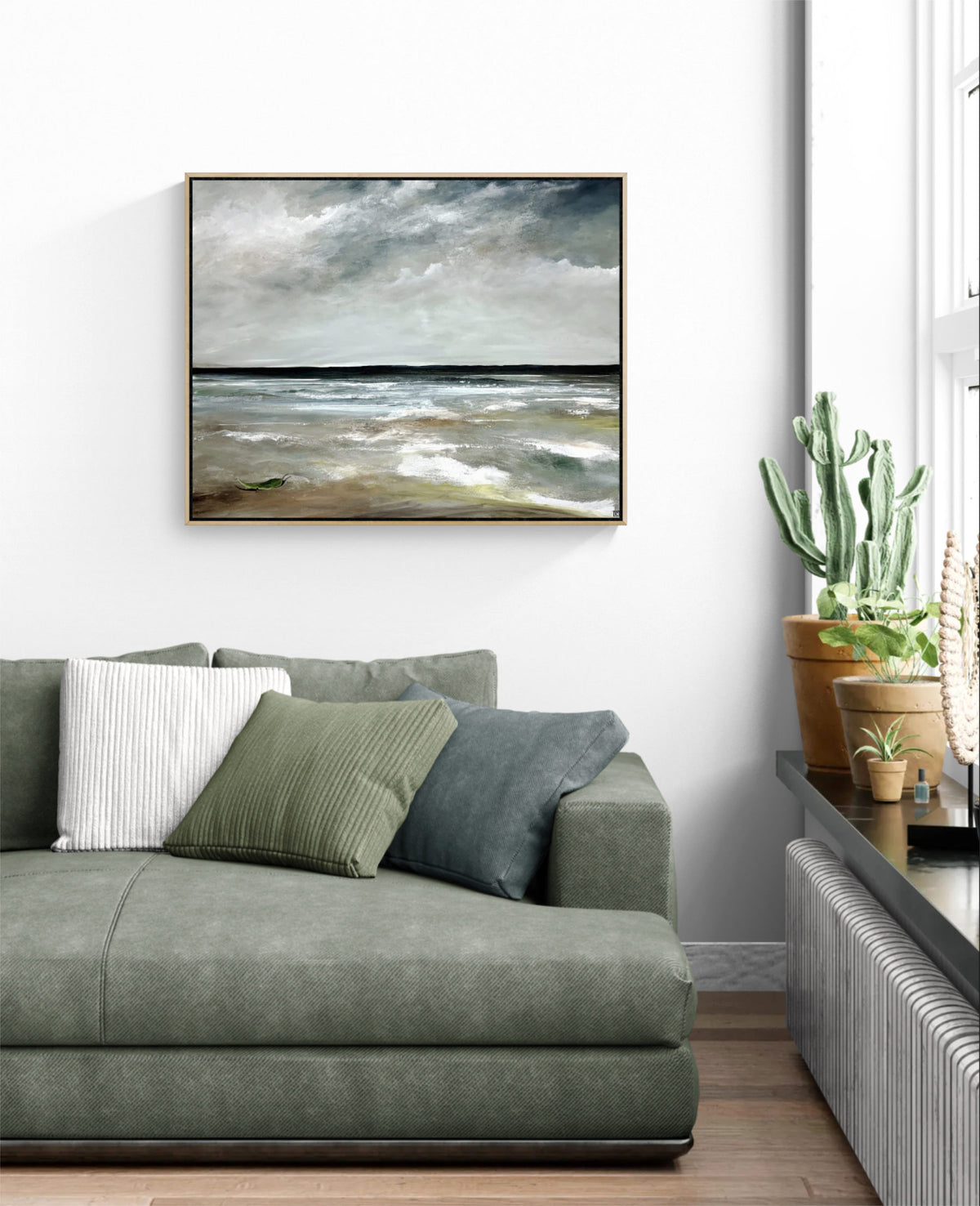 A framed horizontal landscape painting of a stormy sea hangs on a white wall above a green sofa in a modern living room setting.