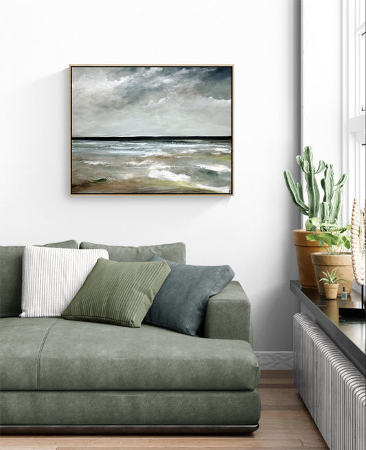 A framed horizontal landscape painting of a stormy sea hangs on a white wall above a green sofa in a modern living room setting.