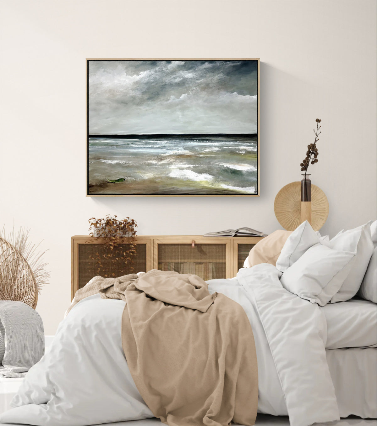 A framed horizontal landscape painting of a stormy sea hangs on a light beige wall above a bed with white linens and a tan throw blanket in a styled bedroom setting.