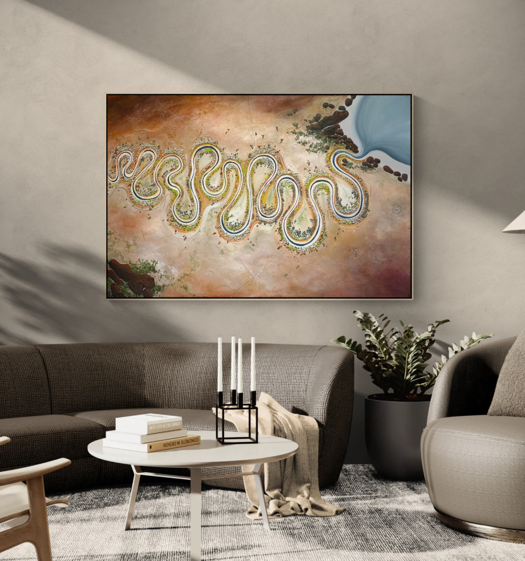 A framed abstract landscape painting featuring a winding river or coastline in blue, green, and gold tones over a warm beige and brown background, displayed on a gray wall above a modern living room setup with a sofa, coffee table, and plant. 180x120cm, acrylic on canvas.