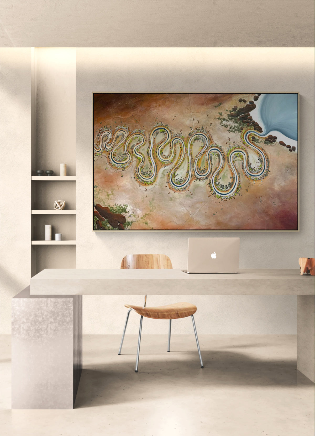 A large framed abstract landscape art print hangs on a light beige wall above a modern concrete desk with a wooden chair and a laptop. The artwork depicts a winding river or coastline in earthy tones with blue water.