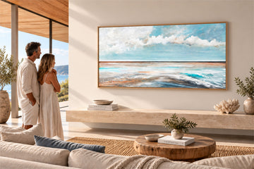 A framed horizontal landscape painting of a beach and ocean under a cloudy sky, displayed on a beige wall in a modern living room. A couple in white robes stands to the left looking at the artwork.