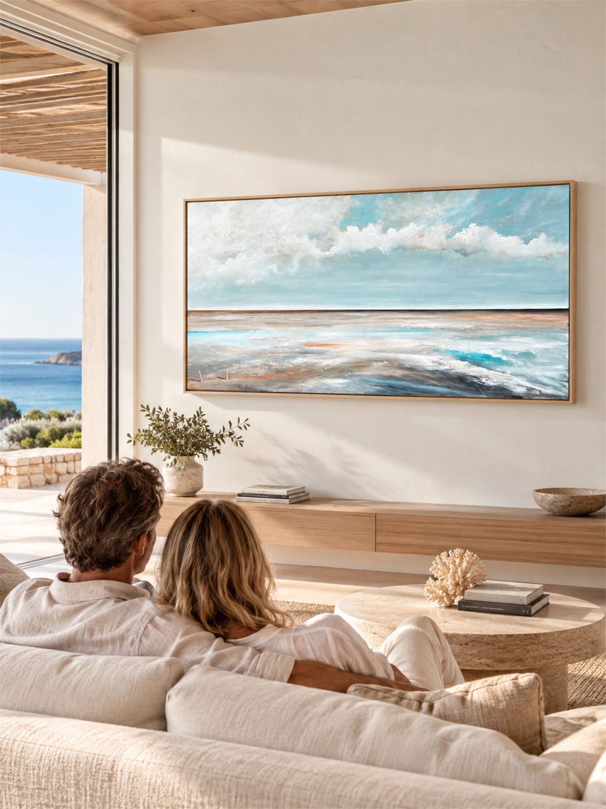 A couple sits on a light beige sofa in a bright, modern room, facing a large framed horizontal painting of a blue ocean and cloudy sky. The room has warm neutral tones, wood ceiling details, and a large window overlooking the sea.