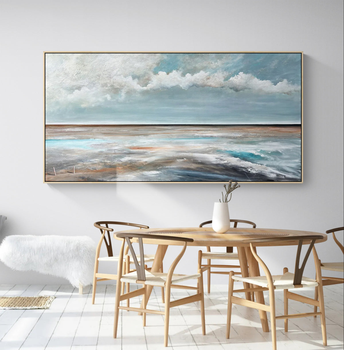 A large framed horizontal landscape painting of a cloudy blue sky over a calm sea, displayed on a white wall above a wooden dining table and chairs in a bright modern room.