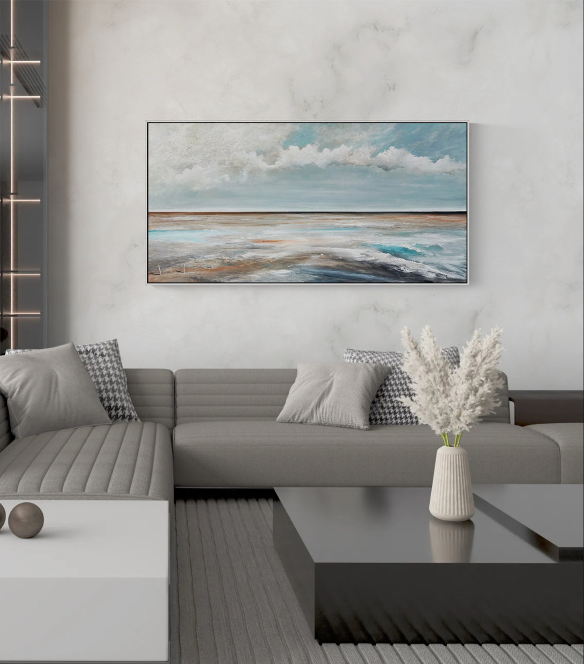 A horizontal landscape painting of a beach and ocean under a cloudy sky, displayed on a light gray marble wall above a modern gray sectional sofa in a living room setting.