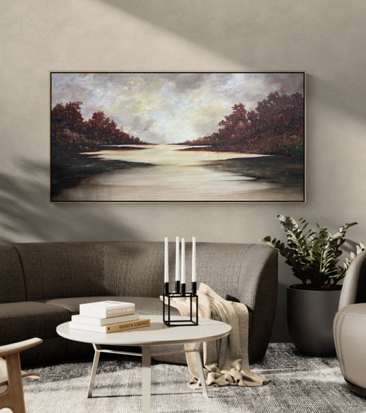A framed landscape painting of a calm river or lake with red-brown trees on both sides and a bright reflective waterway in the center, displayed on a beige wall above a modern gray sofa in a styled living room.