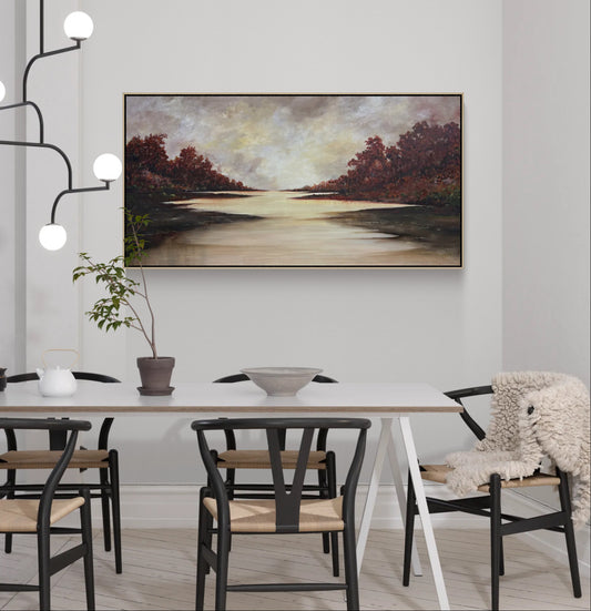 A framed landscape painting of a calm river or lake with autumn trees, displayed on a light gray wall above a dining table in a modern dining room setting.