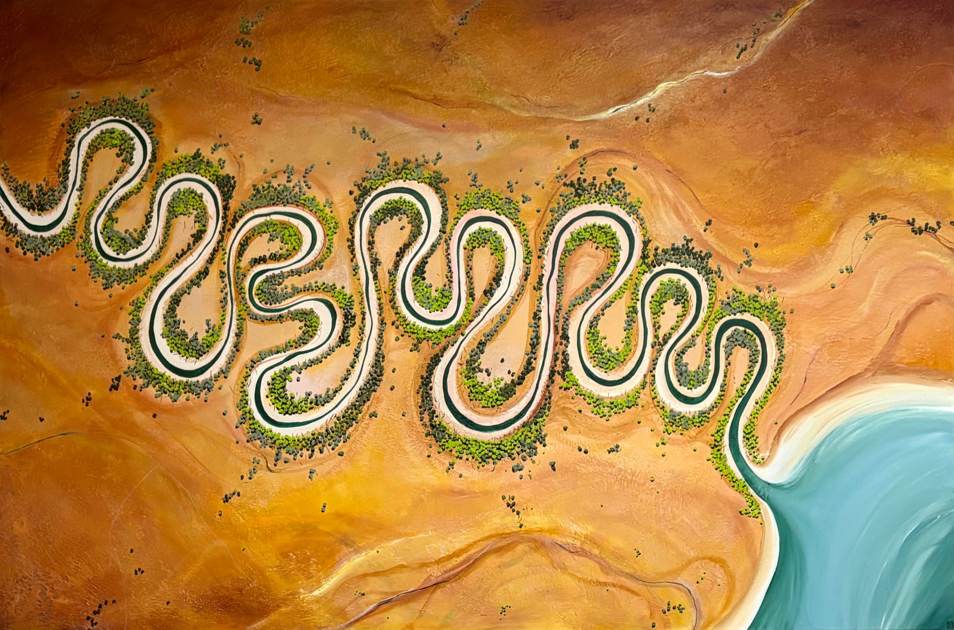 Aerial view of a winding serpentine road cutting through a vast orange desert landscape, with green vegetation lining the road and a turquoise body of water in the lower right corner.