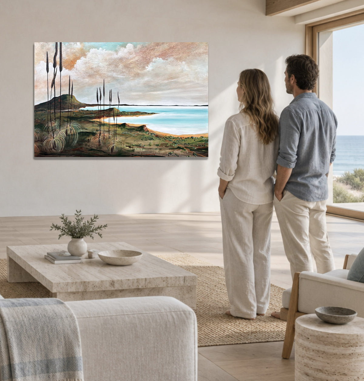 A couple stands in a bright, modern living room looking at a landscape painting of a coastal beach and mountains on the wall.