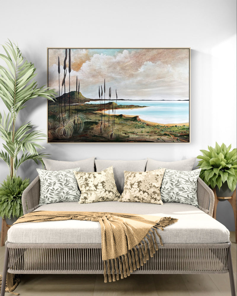 A framed landscape painting of a beach and reeds hangs on a white wall above a light gray daybed with patterned pillows and a beige throw blanket, styled with potted plants on both sides.