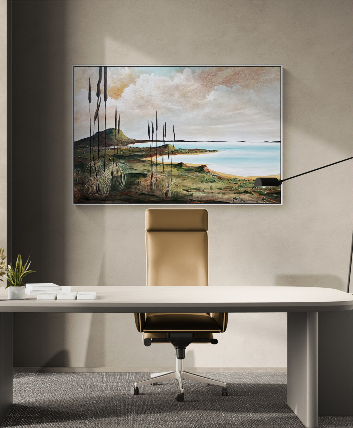 A framed landscape art print depicting a coastal scene with grasses, hills, and water, displayed on a beige wall above a modern white desk and brown leather office chair.