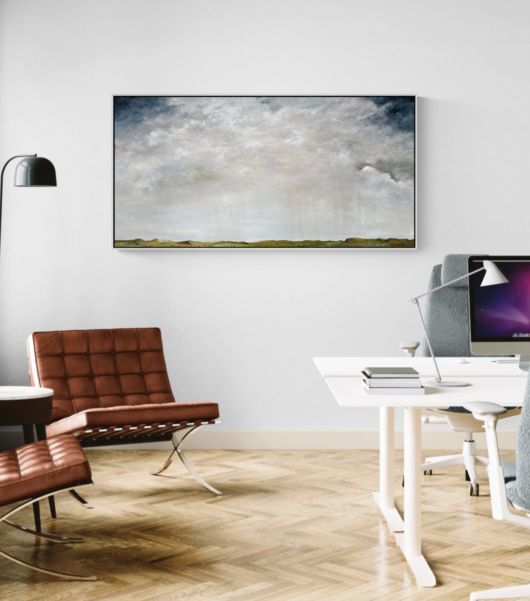 A wide rectangular landscape painting of a cloudy sky over a low green ridge, displayed on a white wall in a modern home office setting with a brown leather chair, desk, and computer.