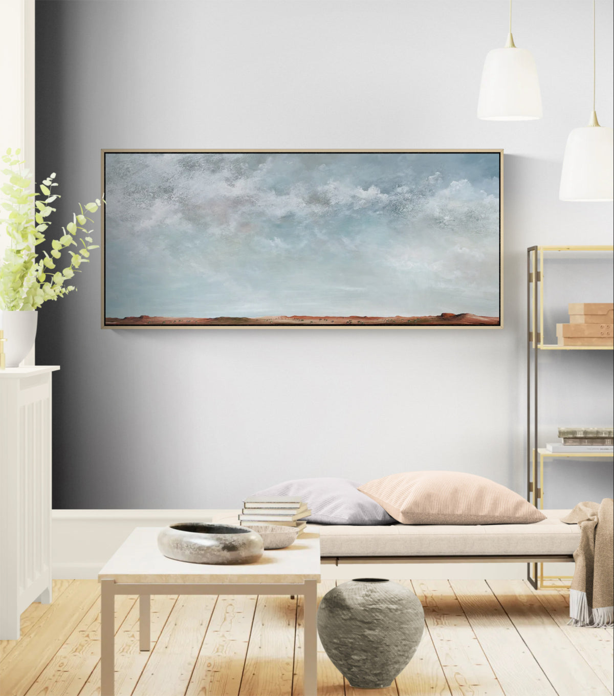 A panoramic landscape painting with a cloudy blue sky and a low sandy horizon, displayed on a gray wall in a modern, sunlit living room setting.