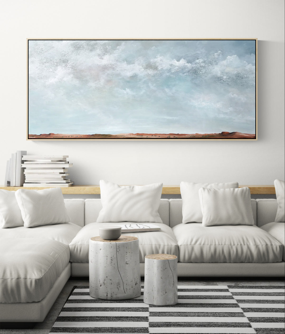 A large horizontal framed painting with an abstract sky scene in soft blue, white, and gray tones with a narrow strip of warm brown land at the bottom, displayed above a light gray sectional sofa in a modern living room setting.