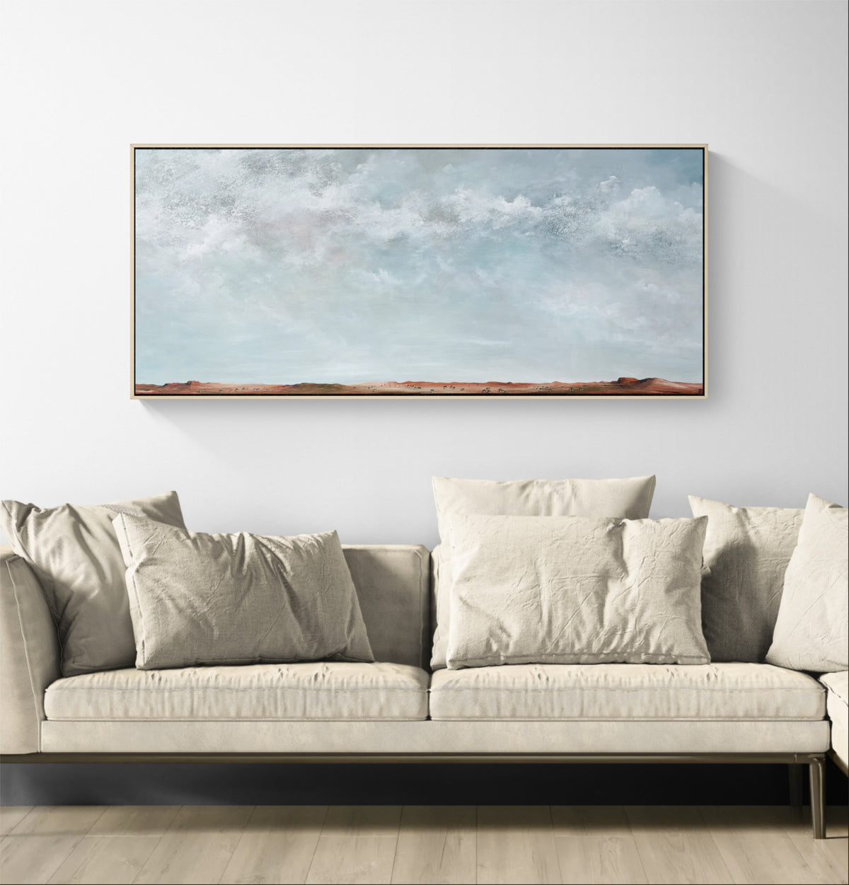A wide rectangular framed landscape painting with a light blue sky and soft clouds hangs above a light beige sofa in a minimalist living room setting.