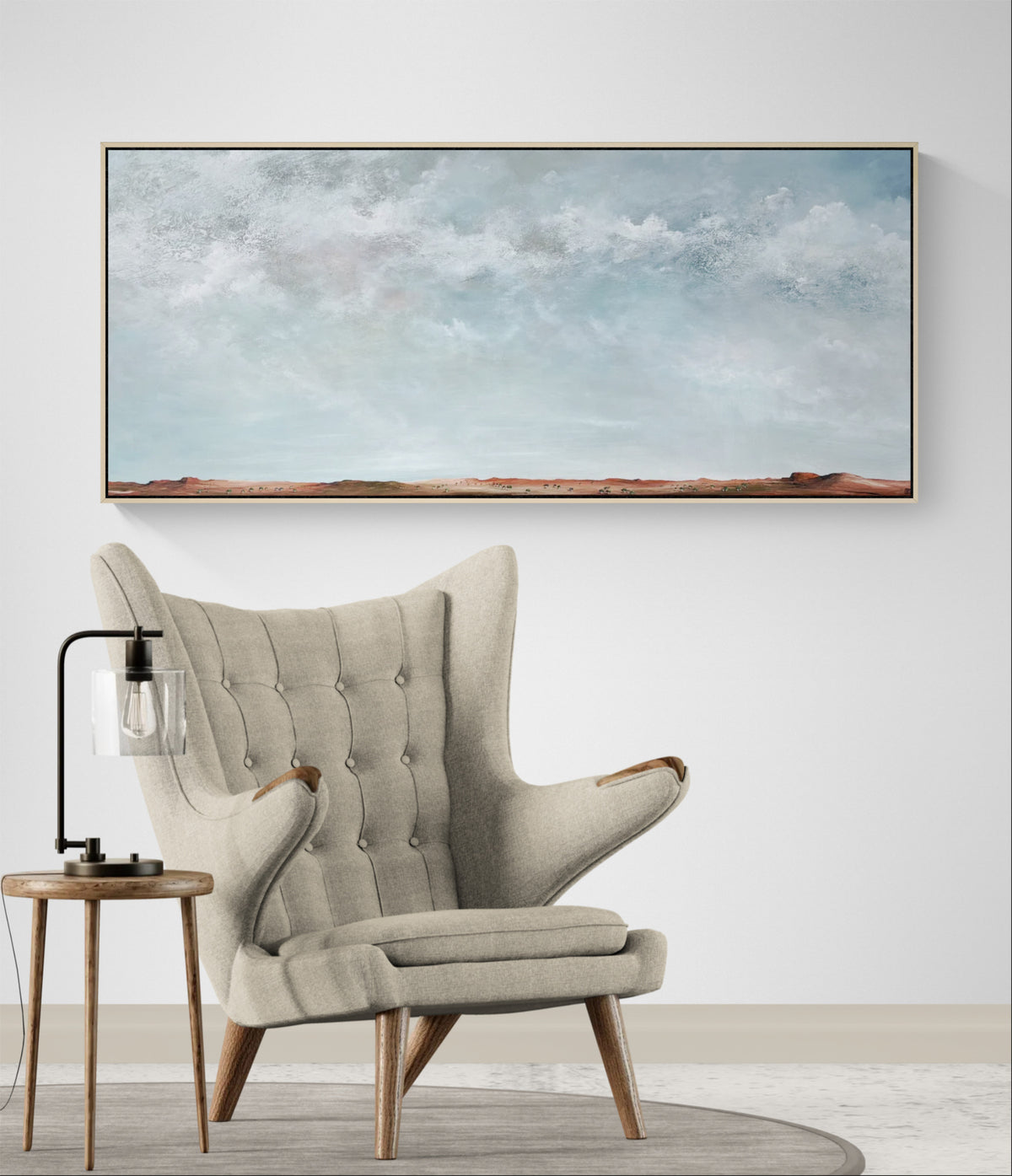 A large panoramic landscape painting with a light blue sky and distant reddish-brown hills, displayed on a white wall above a beige tufted wingback armchair with a small wooden side table and lamp.